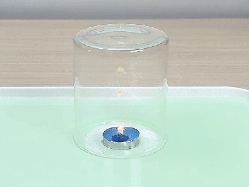 A small, lit tea light candle in a metal holder sits on a white tray filled with a light green liquid. A clear glass cylinder is placed over the candle. The candle flame burns steadily for a short time before gradually diminishing and extinguishing. The green liquid is then drawn up into the glass cylinder. This video demonstrates a classic science experiment illustrating the consumption of oxygen by fire and the resulting change in air pressure.