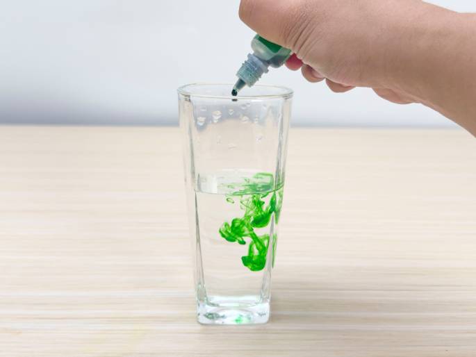 A hand squeezes a small bottle with a dropper tip, dispensing green dye into a clear glass of water on a light wood-grain table. The green dye is visibly dispersing in the water, creating swirling patterns. This image likely depicts a simple science experiment or demonstration illustrating diffusion or solubility.
