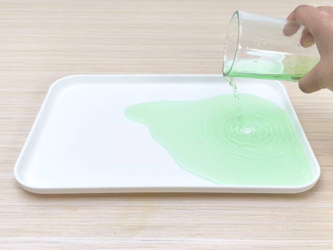 A hand pours light green liquid from a clear glass onto a white rectangular tray resting on a light wood-grain surface. The liquid spreads across the tray, forming ripples and concentric circles where it's being poured. This image likely illustrates a step in a science experiment or demonstration, possibly involving mixing liquids or observing fluid dynamics.