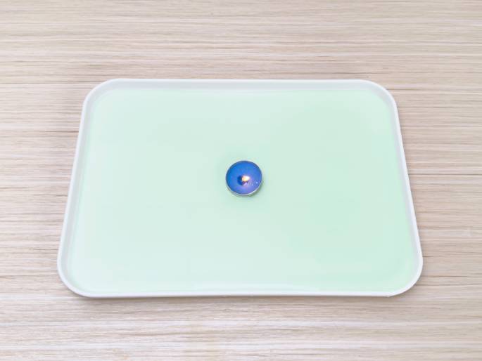 A small, lit blue tea light candle sits in the center of a white rectangular tray filled with a shallow layer of pale green liquid. The tray rests on a light wood-grain table. This setup likely depicts a stage in a science experiment or demonstration, possibly related to combustion, density, or the properties of liquids.