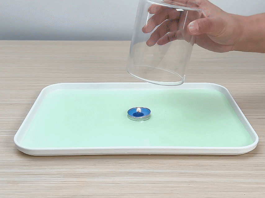 A hand holds a clear glass cylinder above a small, lit blue tea light candle sitting in a shallow pool of pale green liquid within a white rectangular tray. The tray rests on a light wood-grain table. The hand appears to be about to place the glass over the candle, likely as part of a science experiment demonstrating the effects of oxygen depletion on a flame.