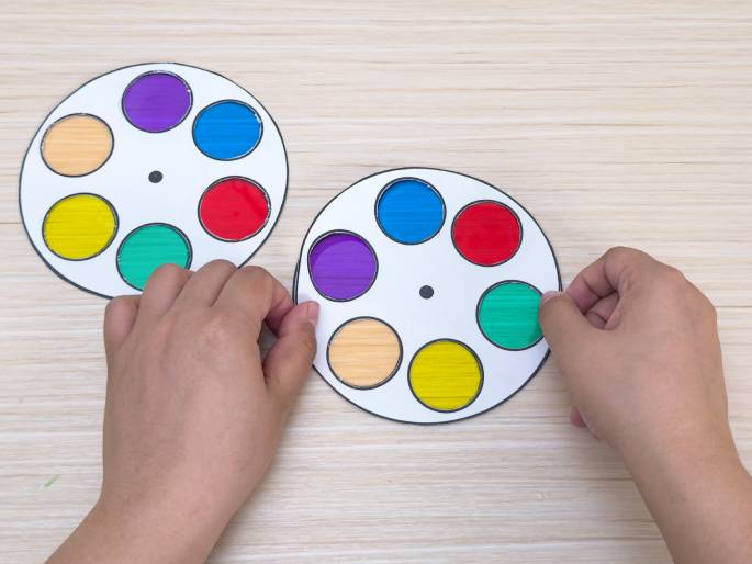 Two hands interact with a pair of color wheels on a light wooden table. The wheels are made of white paper with circular openings filled with translucent colored cellophane. The colors include red, yellow, green, blue, purple, and orange. One wheel is slightly further away, while the closer wheel is being actively turned by one of the hands. This image likely demonstrates a color-mixing activity or an educational toy related to color theory.