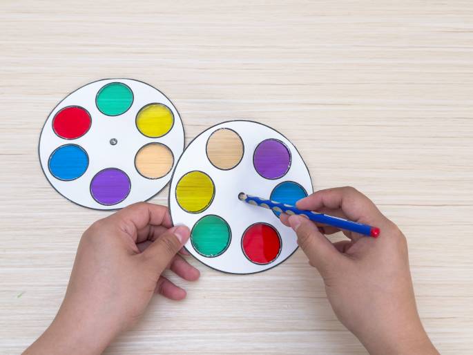 Two hands manipulate two color wheels on a light wooden background. The wheels are made of white paper with circular cutouts covered with translucent colored cellophane. The colors include red, yellow, green, blue, purple, and orange. One hand holds one wheel steady, while the other hand uses a blue pencil to spin the second wheel, presumably to demonstrate color mixing or a similar concept.