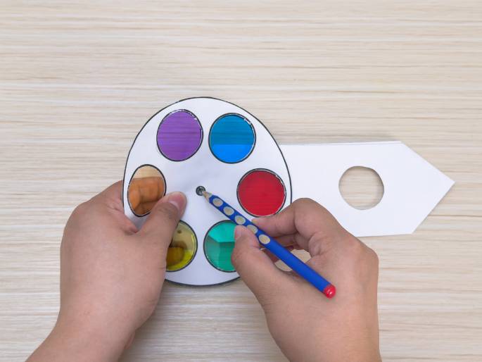 Two hands are shown interacting with a paper color wheel and a separate paper handle on a light wooden surface. The color wheel is white with circular sections of translucent cellophane in various colors, including red, blue, green, purple, orange, and yellow. A blue pencil is being used to spin the color wheel. The handle piece is white and arrow-shaped with a circular hole cut out, designed to be attached to the color wheel. This setup appears to be part of a craft project or educational tool related to color theory and mixing.