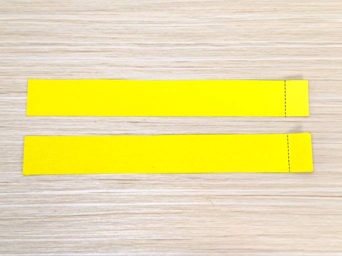 Two identical bright yellow paper strips lie horizontally on a light wood-grained surface. Each strip has a short, dashed, vertical line near its right end, indicating a fold or cut line. These strips likely represent components of a paper craft project.