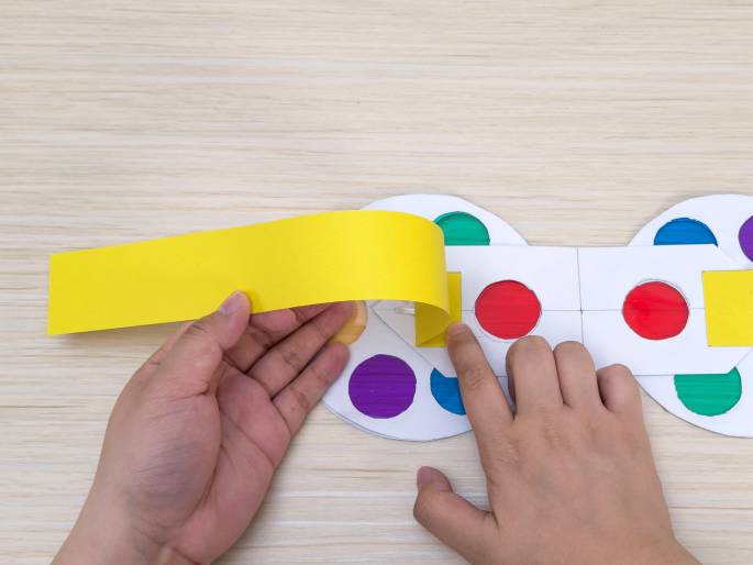 Two hands work on a paper craft project on a light wooden surface. A long, bright yellow strip of paper is being positioned over a white paper construction. The white paper has circular cutouts revealing translucent colored cellophane underneath in red, blue, green, purple, and orange. The yellow strip appears to be part of a mechanism or handle for the color wheels. This image likely depicts the creation of a color-mixing toy or learning tool.