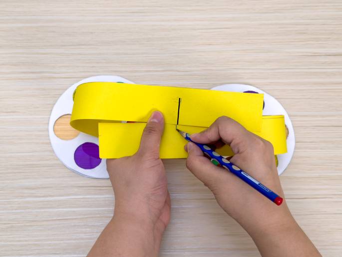 Two hands are constructing a paper craft on a light wooden background. A folded piece of bright yellow paper is being marked with a blue pencil. Partially visible beneath the yellow paper are two white paper color wheels with translucent colored cellophane circles. The colors visible include purple, orange, and bits of green and blue. The pencil is drawing a line, likely a cutting or folding guide, on the yellow paper, which appears to be a handle or mechanism for the color wheels.