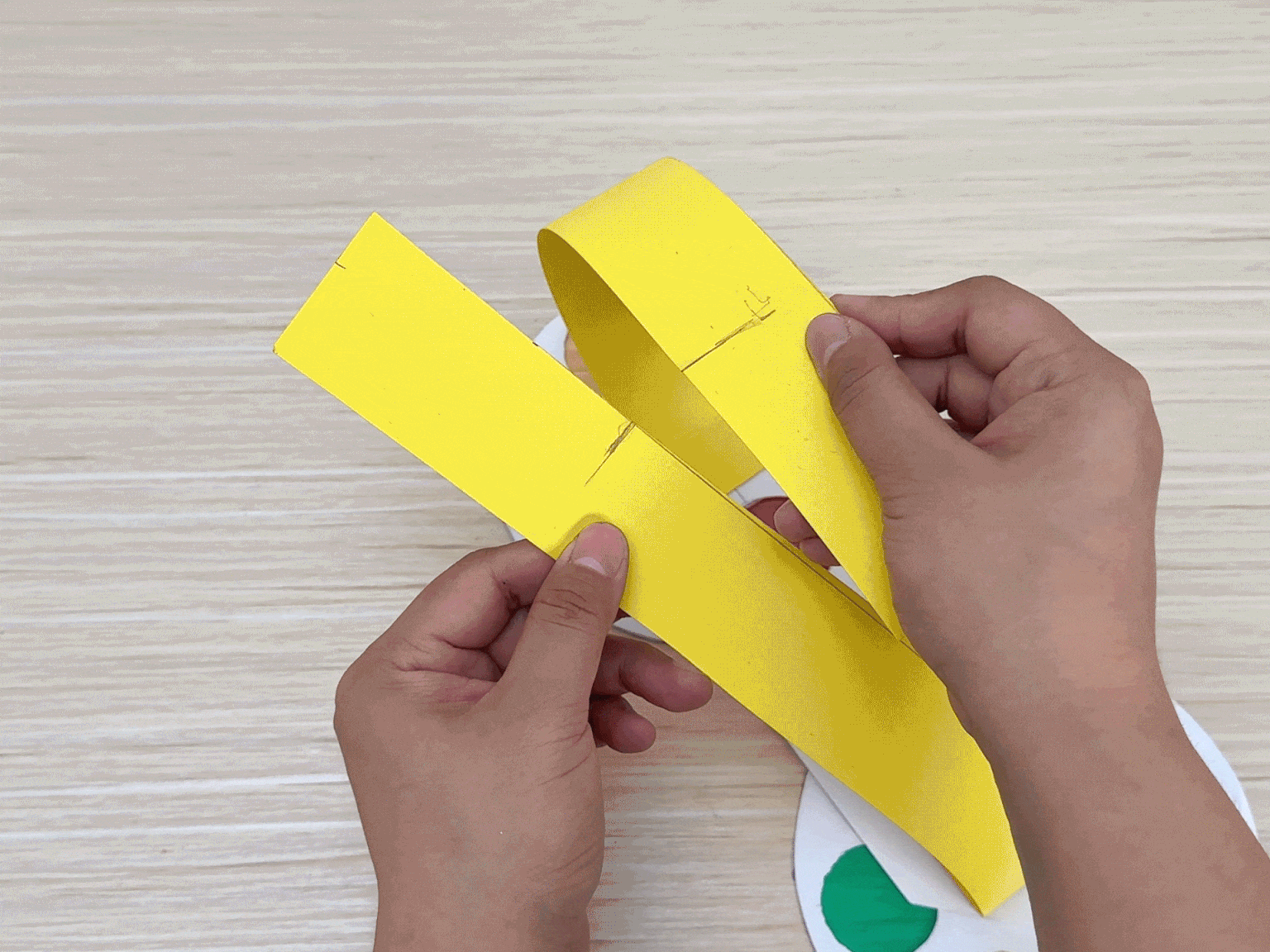 Two hands manipulate pieces of a paper craft on a light wooden surface. They are working with bright yellow strips of paper, one of which is folded. A portion of a white paper circle with a section of green cellophane is visible beneath the yellow strips. Small pencil marks are evident on the yellow paper, likely indicating fold or cut lines. The hands are in the process of assembling or adjusting the yellow components, which appear to be a handle or mechanism for a color wheel device.