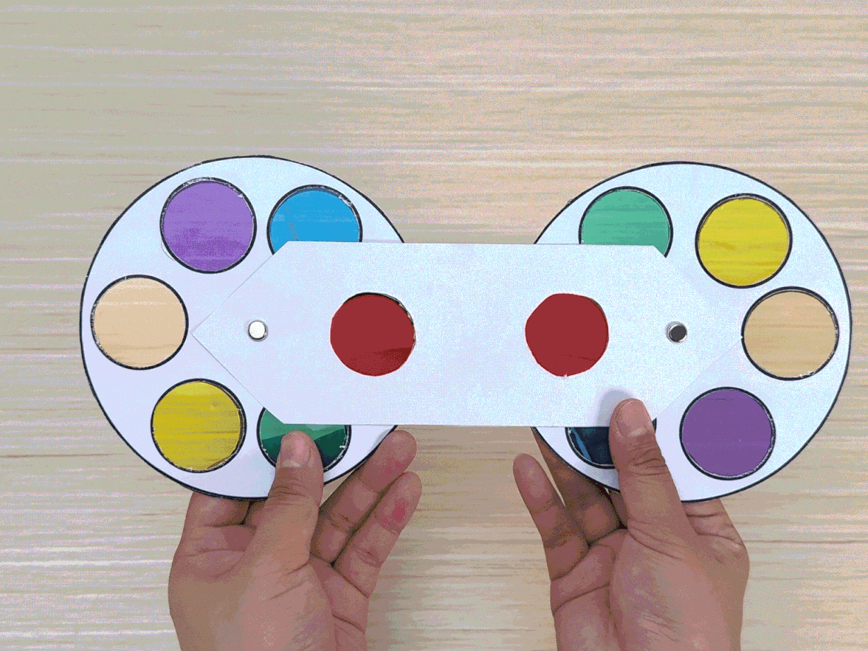 Two hands hold a completed paper color wheel device against a light wooden background. The device consists of two circular color wheels connected by a white paper handle. Each wheel has segments of translucent colored cellophane in red, yellow, green, blue, purple, and peach/orange. The handle has two red circles visible, suggesting a mechanism for viewing color combinations. A small brad or fastener is visible at the center point where the handle joins each wheel. This image showcases the finished color-mixing tool.