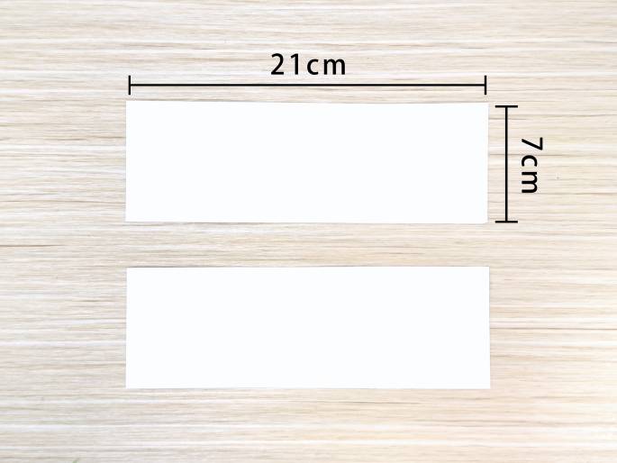 Two blank white rectangular pieces of paper are placed on a light-colored wooden surface. The top rectangle has dimensions marked as 21cm in length and 7cm in width. The bottom rectangle appears to be the same size, though its dimensions are not labeled. This image likely depicts the preparation stage of a paper craft project, showing the size of paper required.