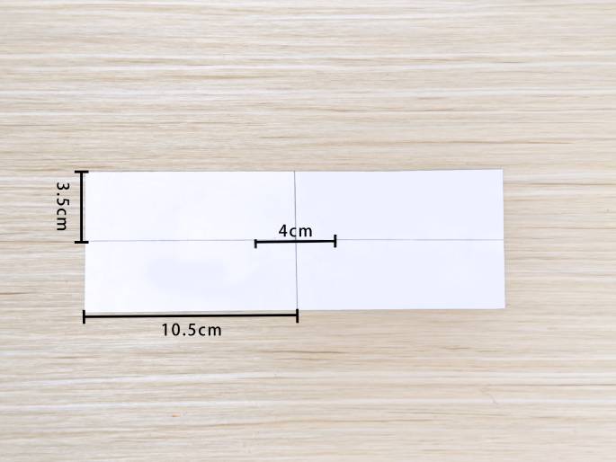 A rectangular piece of white paper rests on a light wood-grain background. Light pencil lines divide the paper into sections. Measurements are marked: 3.5cm for the height, 10.5cm for the width, and a 4cm segment along the horizontal midline. This image likely illustrates the steps in a paper crafting project, showing how to measure and cut the paper.