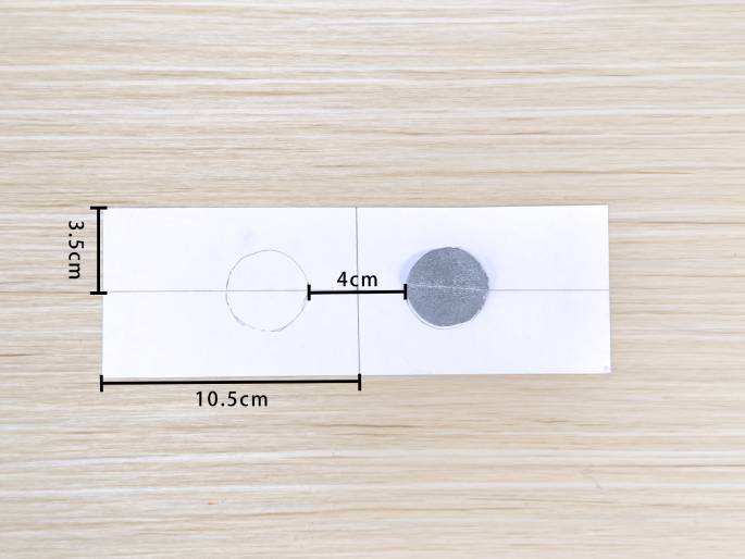 A piece of white paper, marked with dimensions of 10.5cm wide and 3.5cm high, lies on a light wood-grained surface. A pencil-drawn circle is on the left side, and a slightly larger silver foil circle is on the right. A line connects the centers of the circles, marked as 4cm. The image seems to illustrate a step in a craft project, possibly demonstrating the sizing or placement of the foil piece onto the paper.