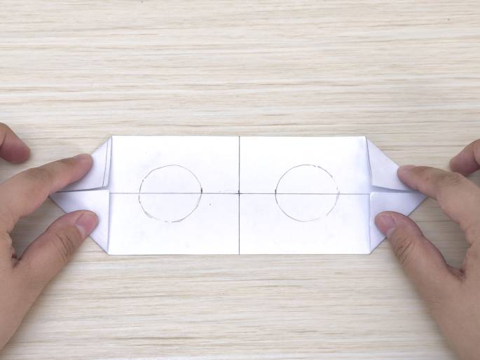 Two hands fold the outer edges of a rectangular piece of white paper inward, towards the center, on a light wooden surface. The paper has two circles drawn on it, one in each half, and a faint vertical line appears in the middle where the folds meet. This image likely demonstrates a step in a paper-folding or origami project.