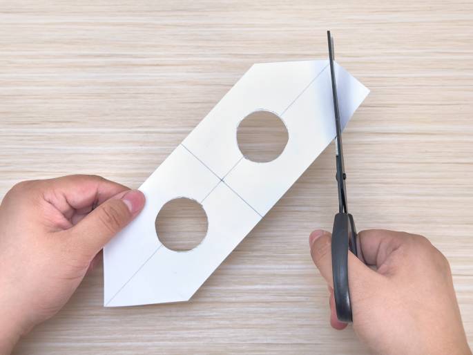 On a light wooden table, two hands manipulate a piece of white paper. The paper has two circular holes cut out and is folded lengthwise with angled edges. One hand holds the paper while the other hand uses scissors to cut off one of the pointed ends. The image suggests a craft project in progress.