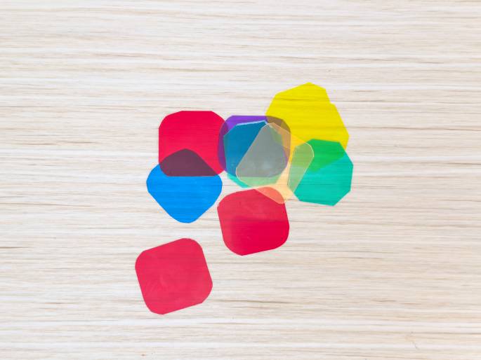 A collection of small, translucent, colored plastic shapes are scattered on a light wooden surface. The shapes are roughly square or octagonal with rounded corners and come in various colors including red, blue, green, yellow, purple, orange, and clear. They appear to be color filters or gels, possibly for photography or lighting.