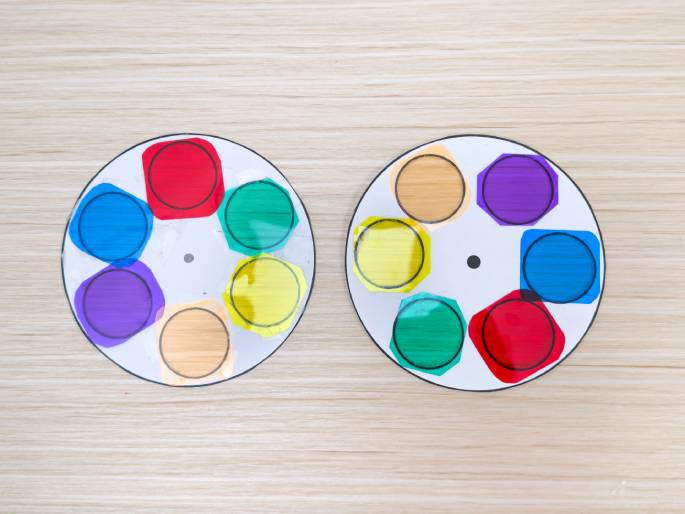 Two circular white paper wheels rest on a light wood-grain surface. Each wheel has translucent colored cellophane shapes attached around the center point, resembling a color wheel. The colors on each wheel include red, yellow, green, blue, purple, and orange. The arrangement of the colors varies slightly between the two wheels. This image likely depicts a learning tool for color theory or a craft project involving color mixing.
