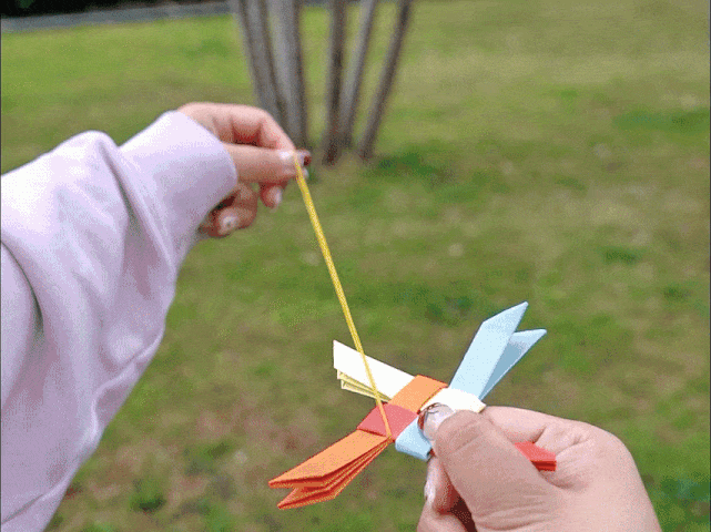 A hand, adorned with a light pink sleeve, aims and launches a small, yellow rubber band outdoors. The rubber band stretches and flies towards a blurred background featuring green grass, trees, and a partially visible building.
