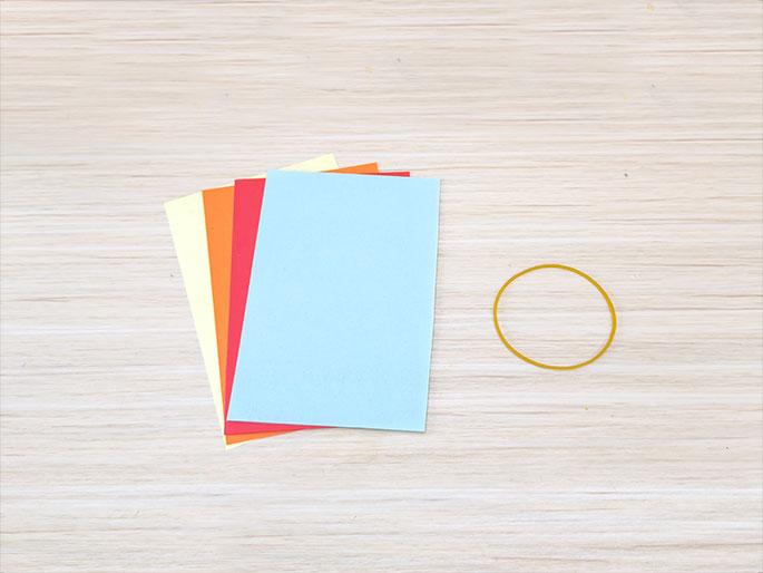 Several rectangular sheets of colorful paper lie on a light wood-grained surface. The visible colors are light blue (on top), red, orange, and pale yellow. A single yellow rubber band is positioned to the right of the paper. The arrangement suggests preparation for a craft project.