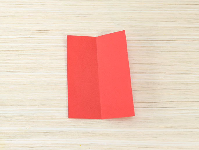 A rectangular piece of bright red paper, folded in half lengthwise, lies on a light wood-grained surface. The fold is visible down the center of the paper.
