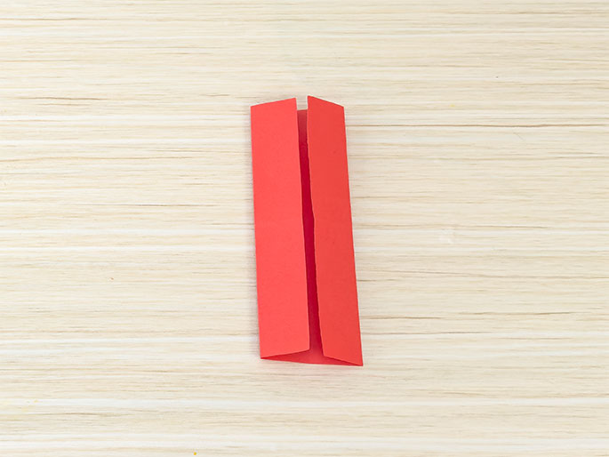 A rectangular piece of red paper, folded in half lengthwise and then in half again widthwise, rests on a light wood-grained surface. The folded edges create a narrow, rectangular shape.