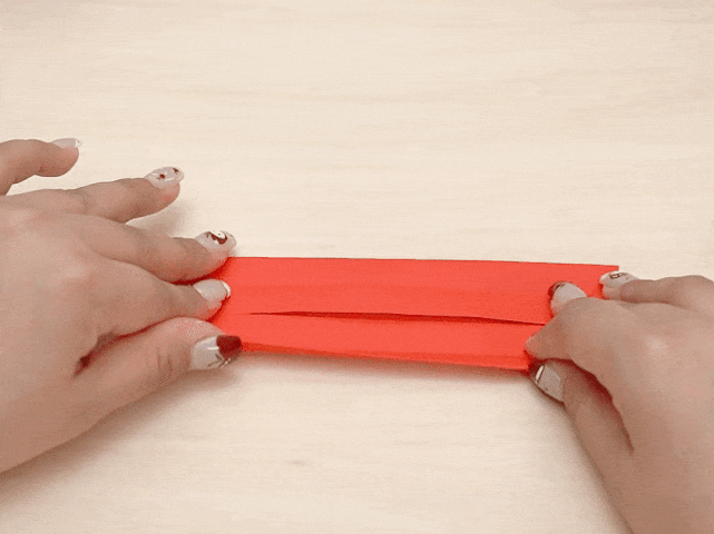 Two hands are folding a rectangular strip of red paper. The paper has already been folded in half lengthwise and is now being folded inward again from the shorter sides, creating a narrower, more compact shape. The hands and paper are positioned on a light wood-grained surface.