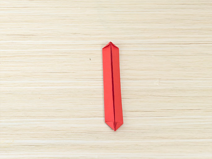 A narrow, folded strip of red paper lies vertically on a light wood-grained surface. Both ends of the strip are folded inwards to create pointed tips, resembling a slim, elongated diamond or a stylized tie.