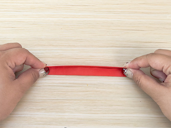 Two hands hold a long, thin strip of red paper against a light wood-grained surface. The paper is folded lengthwise, creating a visible crease down the middle. The hands gently pinch the edges of the paper, appearing to prepare for another fold or manipulation.