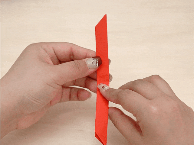 Two hands are manipulating a vertically oriented, narrow strip of red paper against a light wood-grained background. The paper is folded multiple times, and the hands appear to be in the process of making or adjusting a fold near the center of the strip. One end of the paper has a pointed tip.