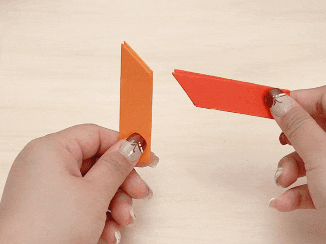 Two hands are shown against a light wood-grained background. One hand holds a small, folded piece of orange paper, while the other hand holds a similar, slightly larger piece of red paper. Both pieces appear to be folded and shaped for an origami project.