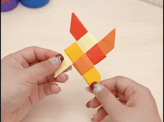 Two hands hold a completed origami ninja star, also known as a shuriken. The star is made of folded paper in shades of orange, red, and yellow, arranged in an interlocking pattern. The points of the star are sharp and defined.