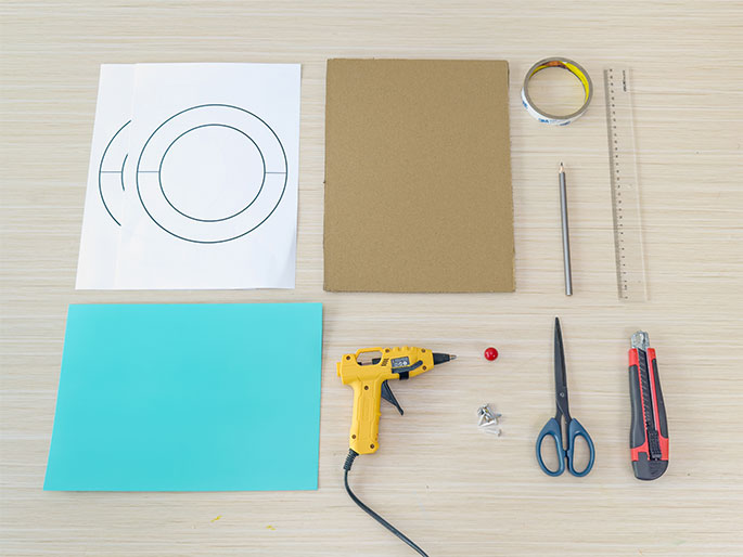 A flat lay arrangement of various craft supplies is displayed on a light wood-grained table. The supplies include: a sheet of white paper featuring concentric circles, a piece of cardboard, a roll of transparent tape, a pencil, a ruler, a sheet of teal paper or cardstock, a yellow hot glue gun, a red bead or pom-pom, several small metal brads, scissors, and a utility knife. These items appear to be assembled for a crafting project.