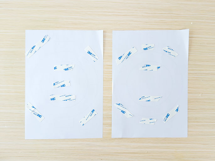 Two sheets of white paper rest on a light wood-grained surface. Several small pieces of 3M double-sided adhesive tape are scattered across both sheets. Some of the tape pieces appear to have been partially used or torn.