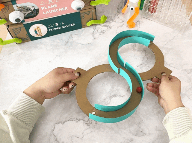 Hands manipulate a figure-eight shaped marble run made from cardboard and teal-colored paper strips. A small red ball is visible within the track of the lower loop. In the background, portions of two boxes labeled PLANE LAUNCHER and FLYING SAUCER are visible, suggesting a crafting or science kit context. The background is a marbled white surface.