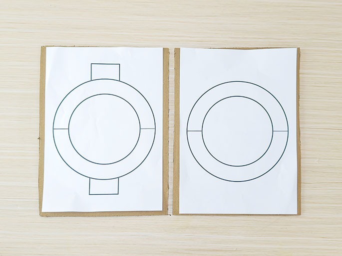 Two pieces of white paper, each with a circular design drawn on them, are placed side-by-side on a light wood-grained surface. The circles are comprised of concentric lines, creating a ring-like shape. The left-hand paper has small rectangular tabs extending from the top and bottom of the circle. The right-hand paper has only the circular design. Both papers appear to be affixed to slightly larger pieces of cardboard.