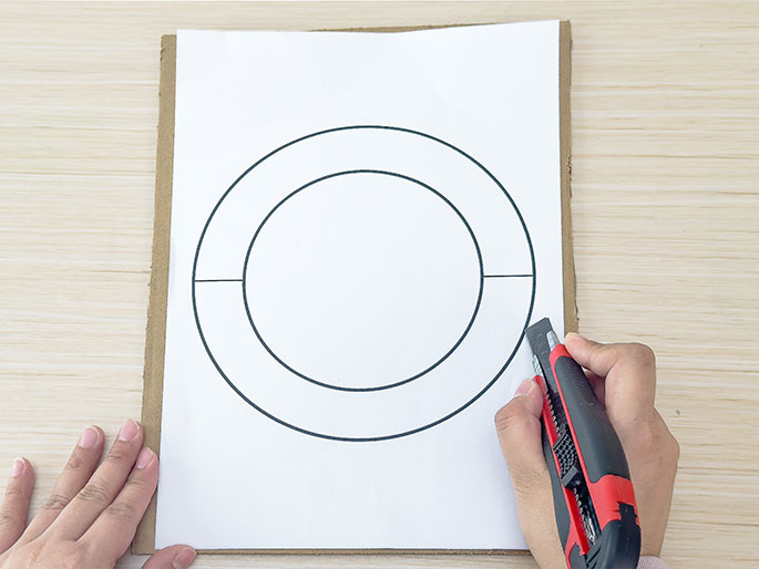 Two hands are shown working on a craft project. One hand holds down a piece of white paper with concentric circles drawn on it, which is placed on top of a piece of cardboard. The other hand holds a red and black utility knife and is beginning to cut along the lines of the innermost circle. The project appears to be taking place on a light wood-grained work surface.
