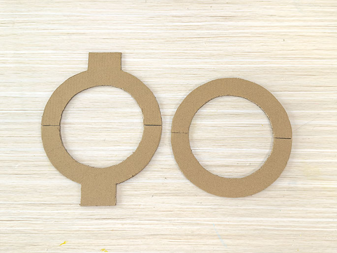 Two cardboard rings rest on a light wood-grained surface. The ring on the left has small rectangular tabs extending from its top and bottom edges. Both rings have a small slit cut into their sides, indicating they were likely cut from a larger piece of cardboard.