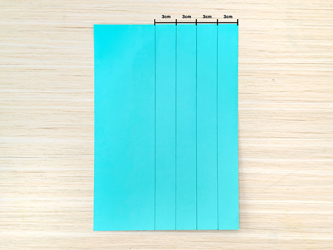 A rectangular piece of teal paper or cardstock lies on a light wood-grained surface. Four vertical lines are drawn on the paper, dividing it into five equal sections. The width of each section is marked as 3cm.