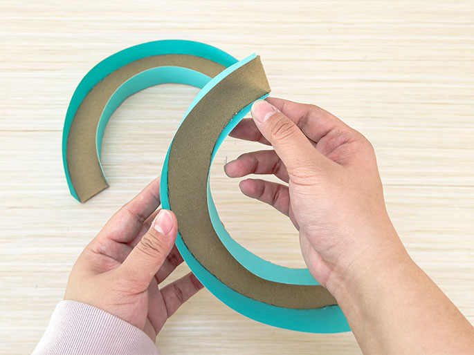 Two hands hold a partially formed teal-colored paper or cardstock ring against a light wood-grained background. The ring is being constructed with a strip of cardboard sandwiched between the teal material. A second, shorter piece of the teal-covered cardboard is visible to the left, suggesting the process of creating the ring. The hands appear to be gently curving the materials.
