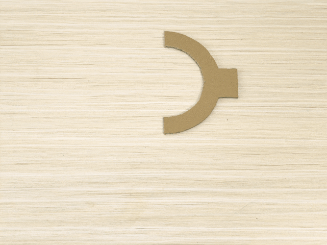 A C-shaped piece of cardboard sits on a light, wood-grained surface. The cardboard piece has a small rectangular tab extending from one end. The majority of the image is the textured background.