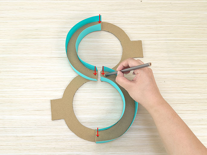 On a light wood-grained surface, two teal-colored paper strips are being attached to two cardboard rings to create a figure-eight shape. One hand holds a pencil and is marking the point where the teal strip meets the cardboard ring on the lower loop of the eight. Red arrows indicate three points of connection between the teal strips and the cardboard rings.