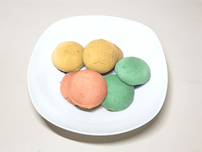 A white, slightly square-shaped plate holds a small assortment of colorful, round cookies. There are two pale yellow cookies, two light orange/pink cookies, and two pale green cookies. They appear to be small and possibly homemade. The plate sits on a plain off-white surface.