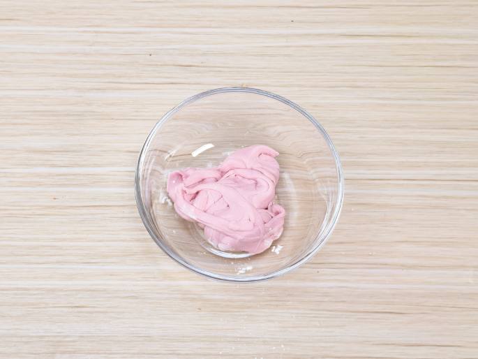 A clear glass bowl, placed on a light wood-grained table, contains a mass of light pink, dough-like or putty-like material. The material appears to be slightly folded and textured. A few specks of white powder are visible in the bottom of the bowl and on the table near the bowl, possibly indicating a dry ingredient used in the mixture. This image likely depicts homemade playdough, slime, or a similar substance.