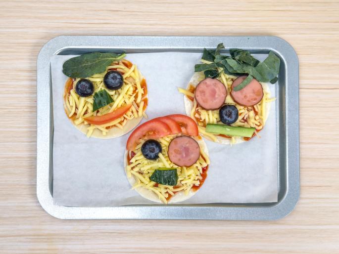 Three small pizzas, decorated to resemble faces, sit on a baking sheet lined with parchment paper. The pizzas have a tomato sauce base and are topped with shredded cheese. Each face uses different toppings for features: one has spinach hair, cucumber noses, tomato mouths, and blueberry eyes; another substitutes sliced sausage for the spinach and tomato; and the last uses a combination of sausage, tomato, and cucumber along with the blueberries and cheese. The baking sheet rests on a light wooden table. These pizzas are clearly designed to be fun and appealing to children.