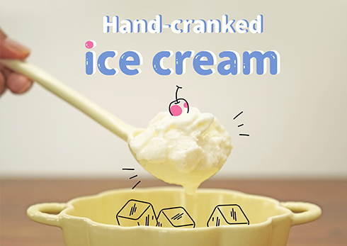 The image cheerfully promotes a Hand-cranked ice cream recipe. A pale yellow bowl, seemingly containing ice cubes, sits in the foreground. Above it, a scoop of creamy, white ice cream, topped with a cartoon cherry, hangs from a pale yellow spoon. The background is softly blurred, focusing attention on the delicious-looking ice cream. The overall style is light-hearted and appealing, suggesting a simple and enjoyable method of making homemade ice cream.