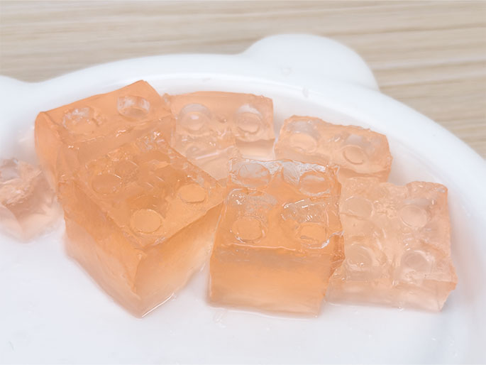 Several pieces of light orange, translucent jelly, molded in the shape of building blocks (similar to LEGO bricks), are arranged on a white plate. The plate has small, rounded, ear-like protrusions, suggesting it's designed for a child. A small amount of clear liquid surrounds the jelly pieces. The image evokes a playful and refreshing treat.