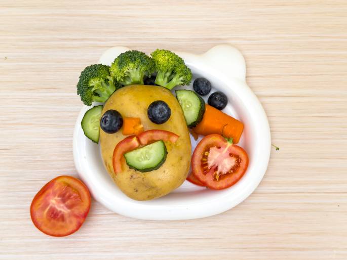 A whimsical face is created using a potato and other vegetables arranged on a white, bear-shaped dish on a light wood-grained table. The potato forms the head, with blueberries for eyes, a small piece of carrot for a nose, and sliced tomatoes and cucumber forming a smiling mouth. Broccoli florets create a hair-like effect on top of the potato. A sliced tomato sits beside the dish. This playful arrangement is likely designed to make healthy eating more appealing, especially for children.