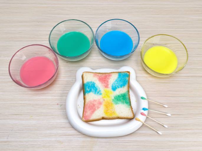 A slice of white bread, decorated with colorful milk paint, sits on a white, bear-shaped dish on a light wood-grained table. The bread is adorned with a radiating design in pink, blue, yellow, and green. Small glass bowls containing the leftover colored milk (pink, green, blue, and yellow) are positioned above the bread. Three cotton swabs, each with a dab of colored milk paint, lie beside the dish, suggesting they were used as painting tools. This image likely depicts a fun food activity for children.