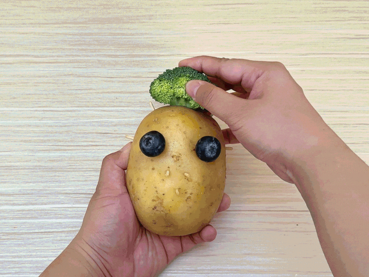 Two hands create a face on a potato using other food items. Broccoli florets are attached to the top of the potato with toothpicks to resemble hair. Two blueberries are secured as eyes, a slice of cucumber forms a smiling mouth, and small pieces of carrot create a nose and ears. The hands arrange and adjust the features on the light wooden table surface. This video appears to demonstrate a fun and creative activity, possibly aimed at children, for making food art.