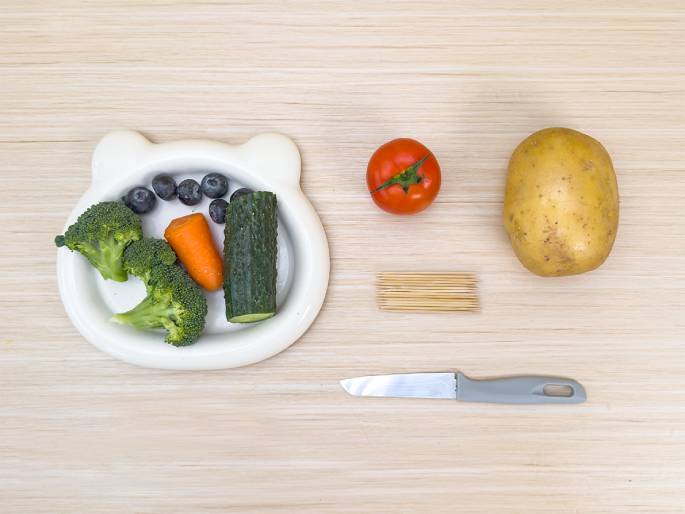 An assortment of food items and utensils are arranged on a light wood-grained table, likely for preparing a meal or snack. A white bear-shaped dish contains broccoli florets, blueberries, a small carrot, and a section of cucumber. Separate from the dish are a single tomato, a raw potato, a bundle of wooden toothpicks, and a small paring knife with a gray handle. The image suggests the ingredients and tools for a healthy and simple food preparation.