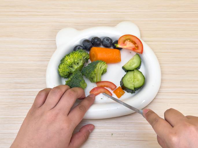 Two hands interact with a white, bear-shaped dish containing chopped vegetables and blueberries on a light wood-grained table. The left hand steadies the dish while the right hand uses a small paring knife to cut a piece of tomato. The dish contains broccoli florets, blueberries, a small, whole carrot, sliced cucumbers, and pieces of a chopped tomato. The scene suggests food preparation, likely for a healthy snack or meal.